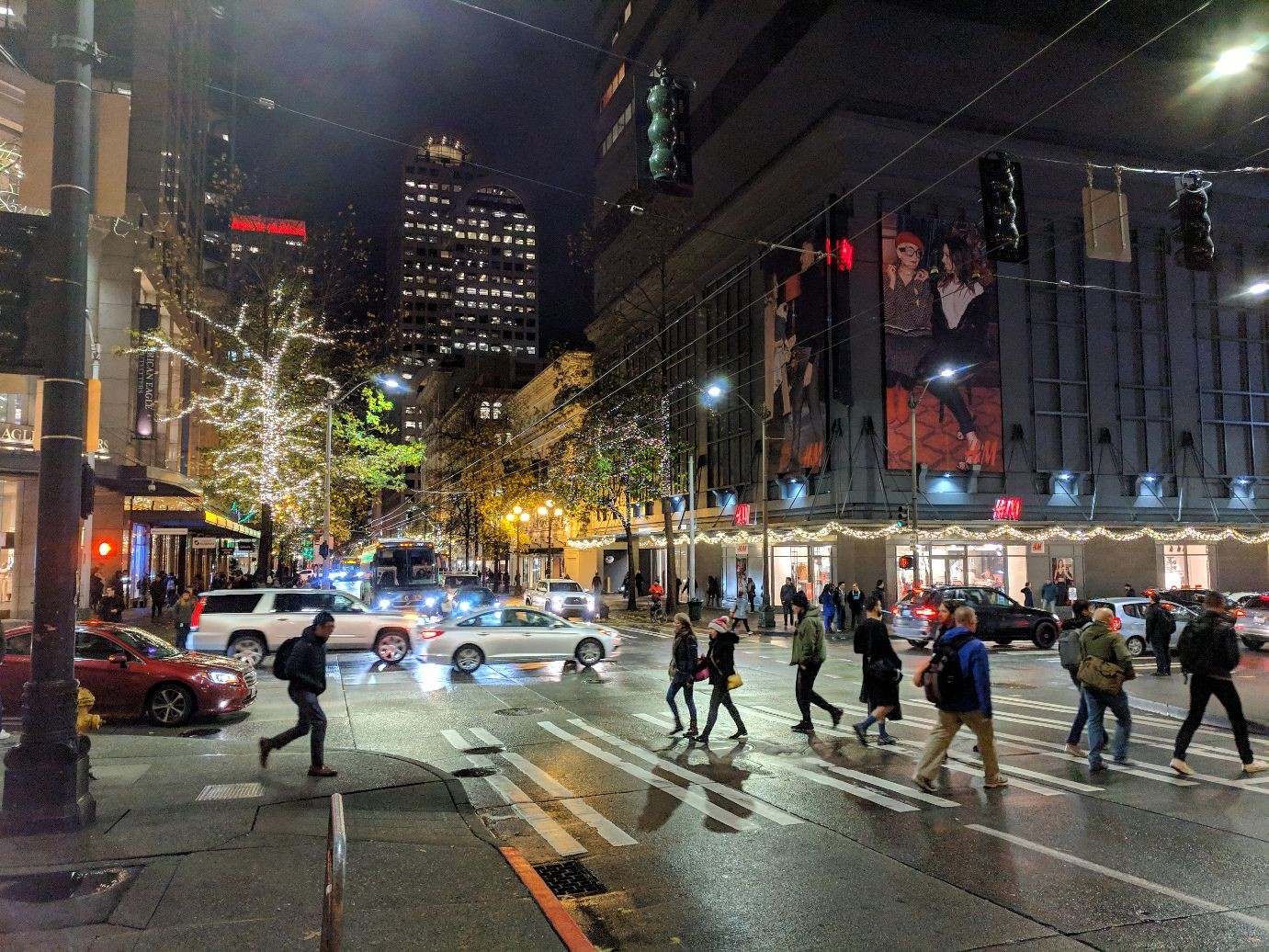 a busy nighttime cityscape to. people are crossing a crosswalk, and trees are lit up with lights.