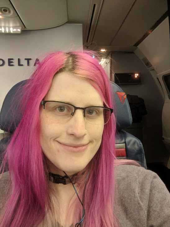 a smiling pink-haired girl on an airplane. She is wearing glasses.