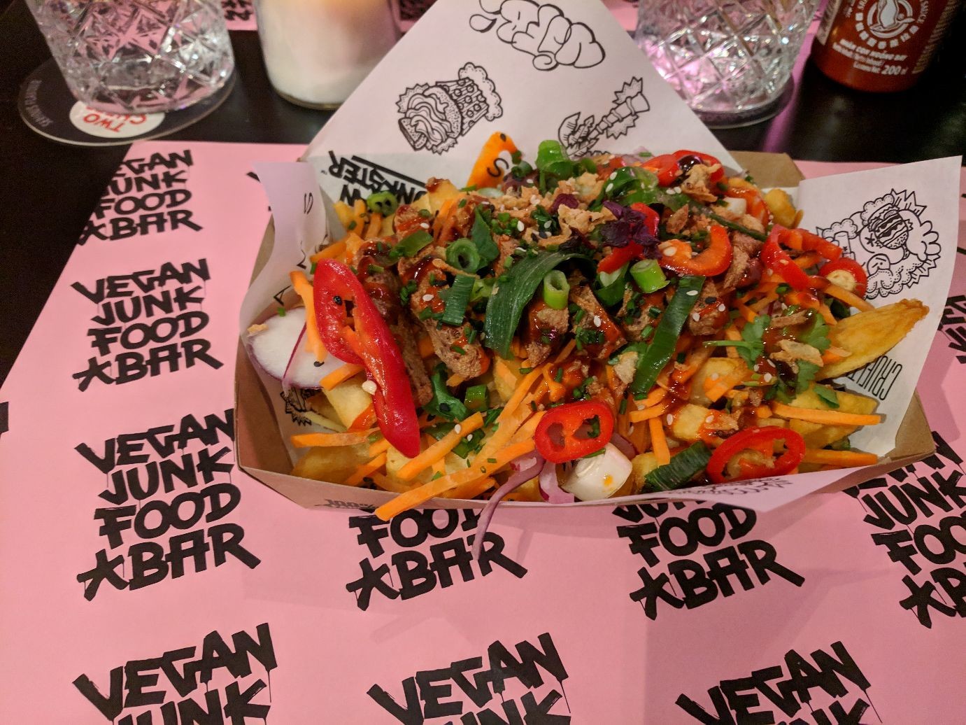 a giant pile of fries covered in sauces and veggies on a paper that says VEGAN JUNK FOOD BAR