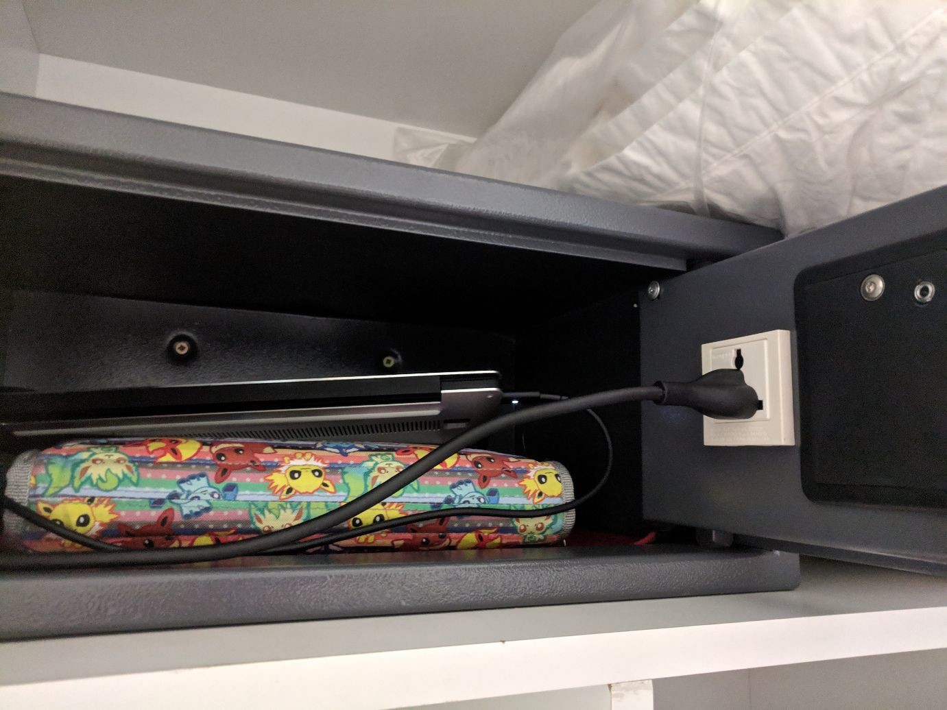 a hotel safe with a power socket in the door. a laptop power cable is plugged into it, and a laptop rests inside propped up on a Switch case.