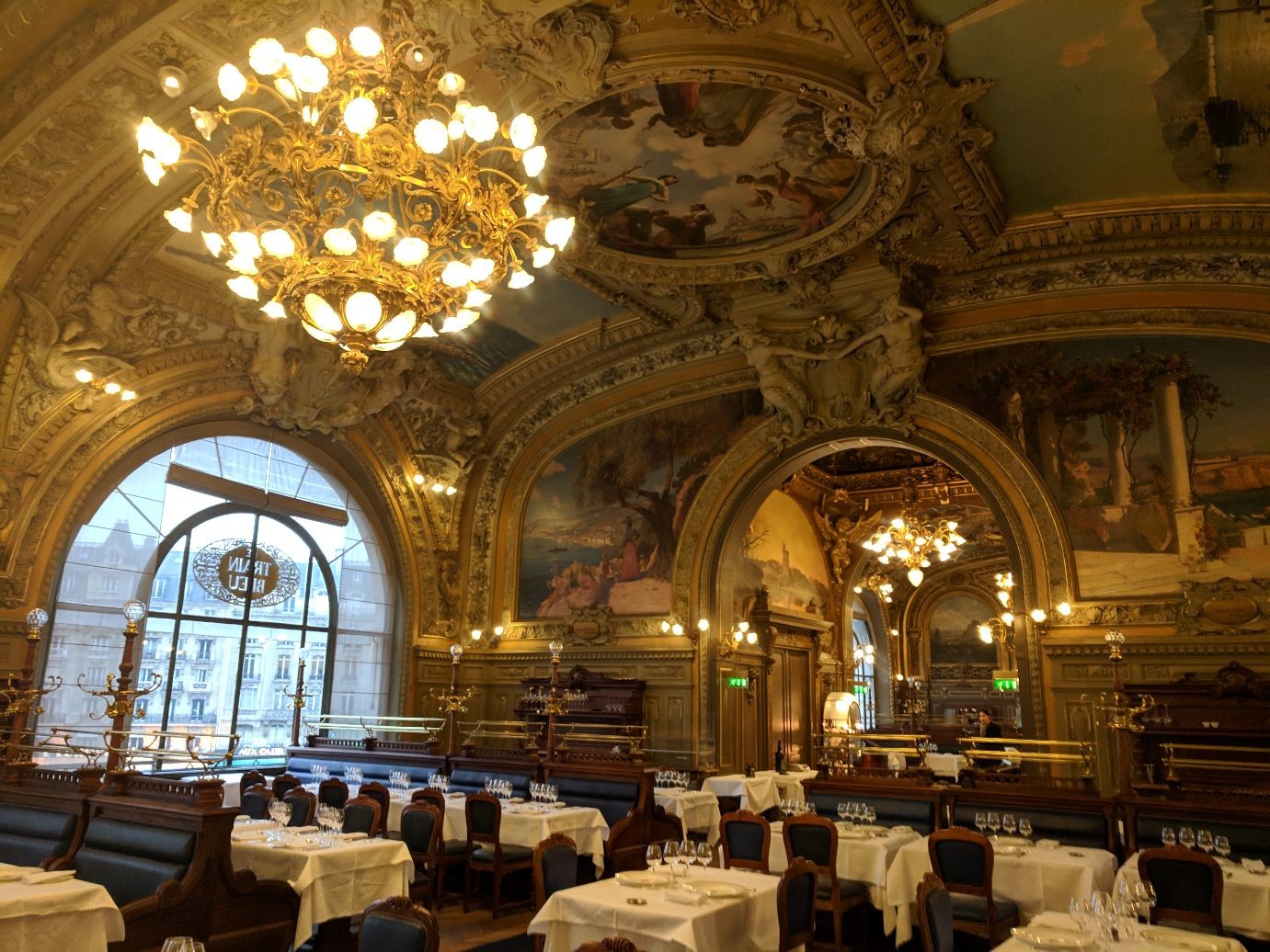 a very fancy restaurant with oil murals on the ceiling interspersed with elaborate stonework and chandeliers. below, tables are set with white tableclothes