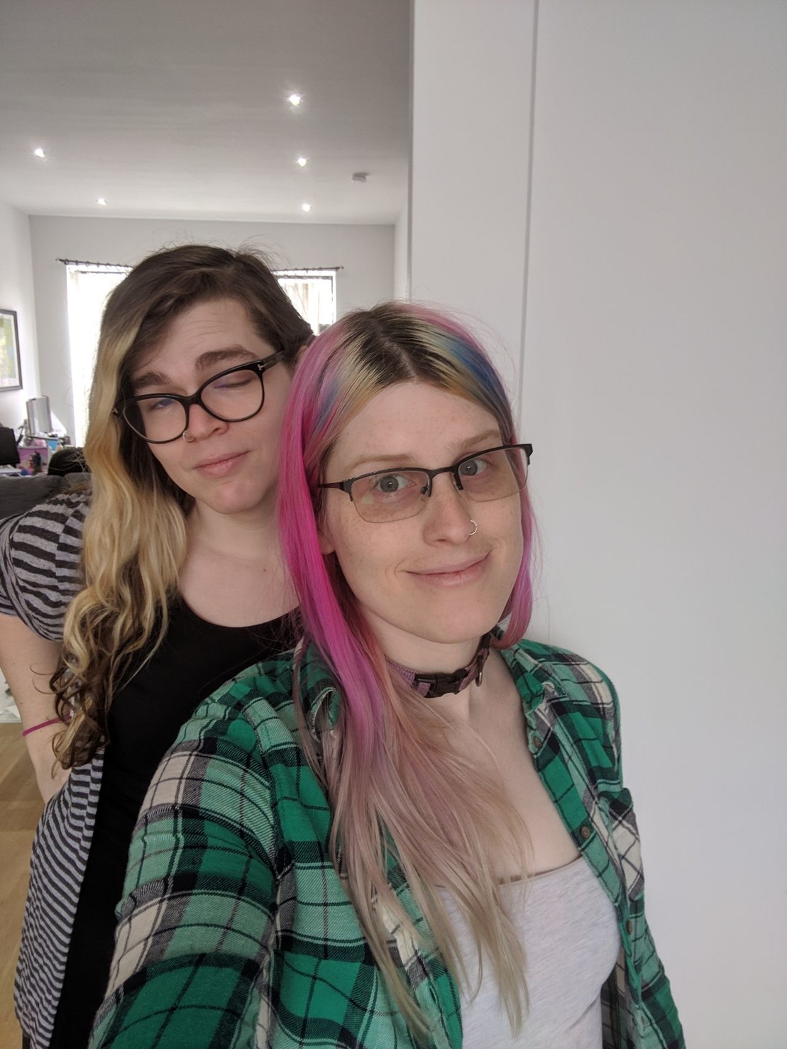 a girl with pink hair in a plaid green top and a white Cami, being photo bombed by a tall lady in a grey cardigan