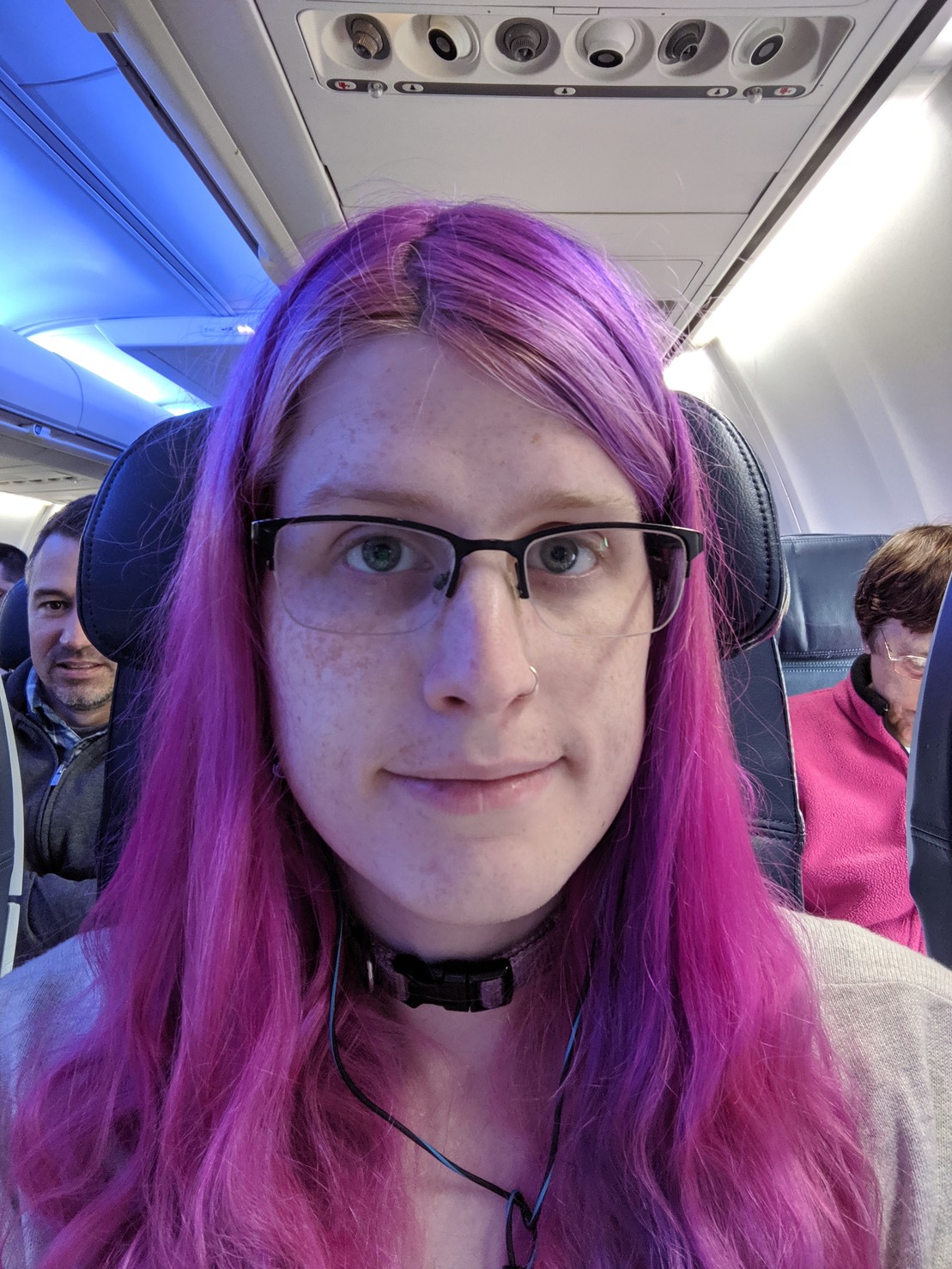 a selfie of me on an airline, smiling weakly. the lighting is blue, my hair is still pink