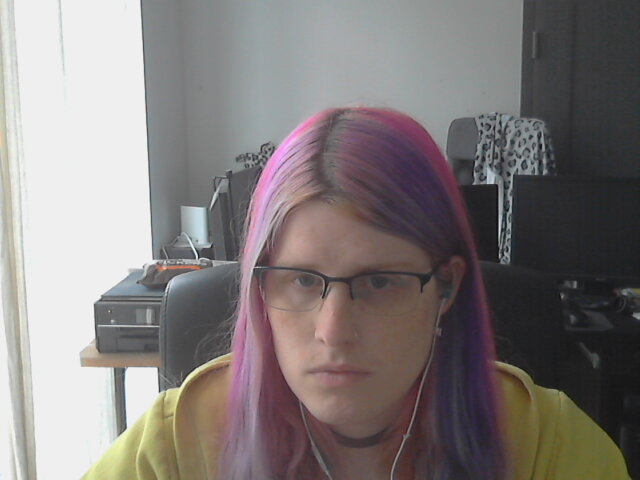 a girl with pink hair and a yellow hoodie in front of a busy background of computers 