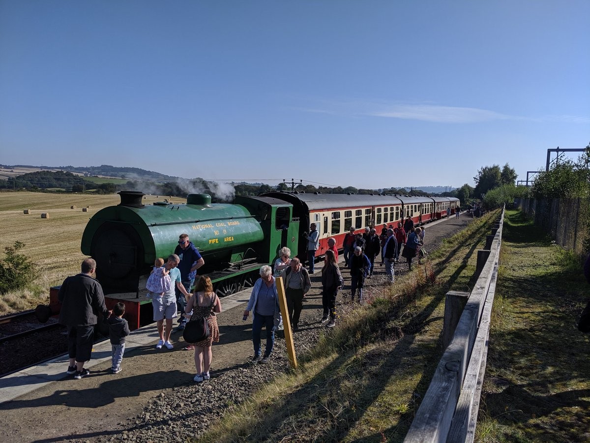 The same green engine coupled to a train of cream and orange passenger carriages. People are milling on a platform