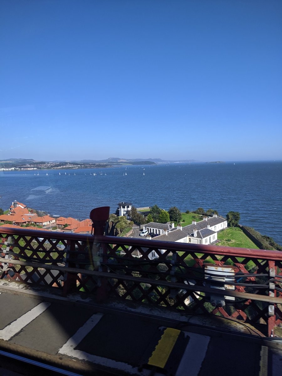 A view off a bridge with large mansion houses in the foreground, and a deep blue ocean dotted with sailboats in the background.