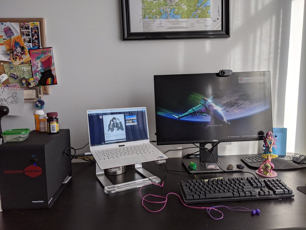 A large black box, a white laptop, and a large monitor showing a spaceship on a desk decorated with a few knicknacks