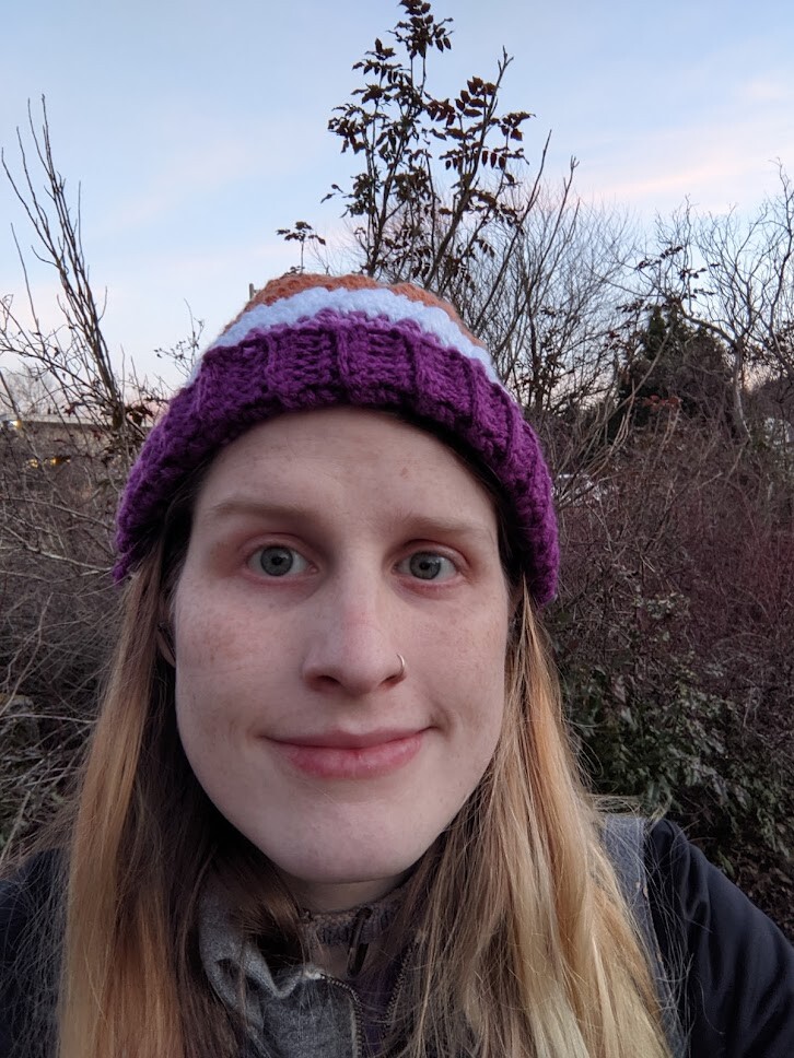 A selfie of me looking at the camera. The background is some bushes, and my hat is Lesbian Pride colours