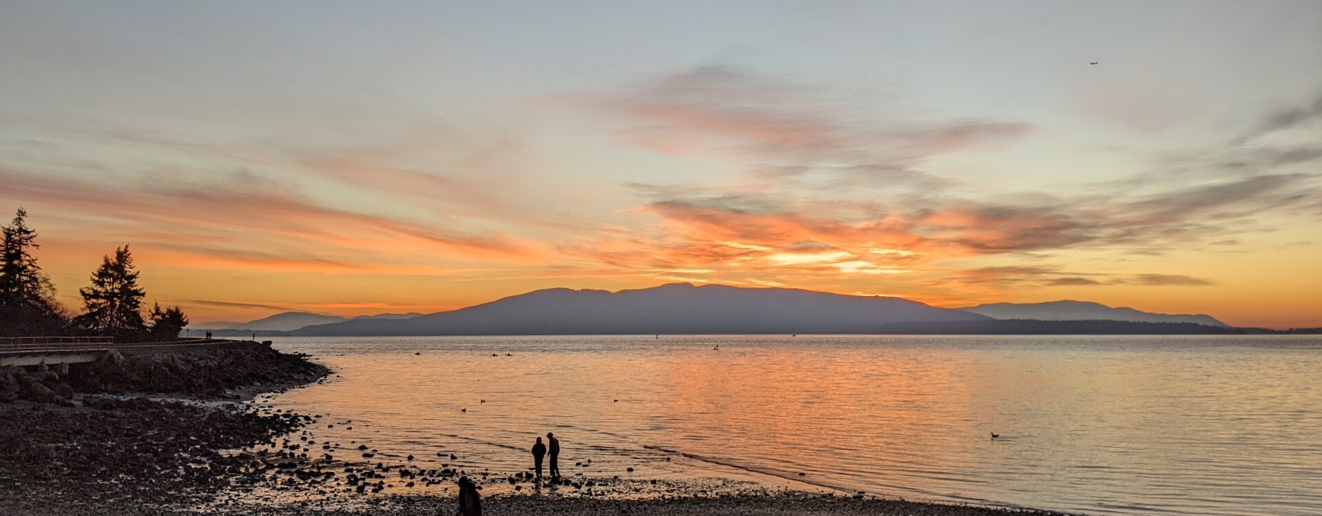 A very orange sunset out over a bay. There's mountains in the distance, and silhouettes of people in the foreground. The orange sunset is reflecting delicately off the water.