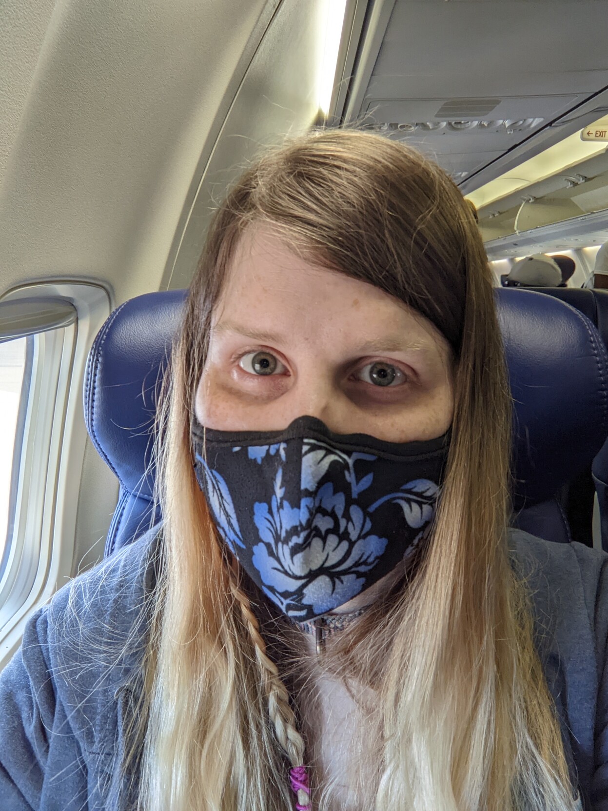 A smiling woman with messy brown and blonde hair and a single thin braid wearing a blue and black mask on an airplane
