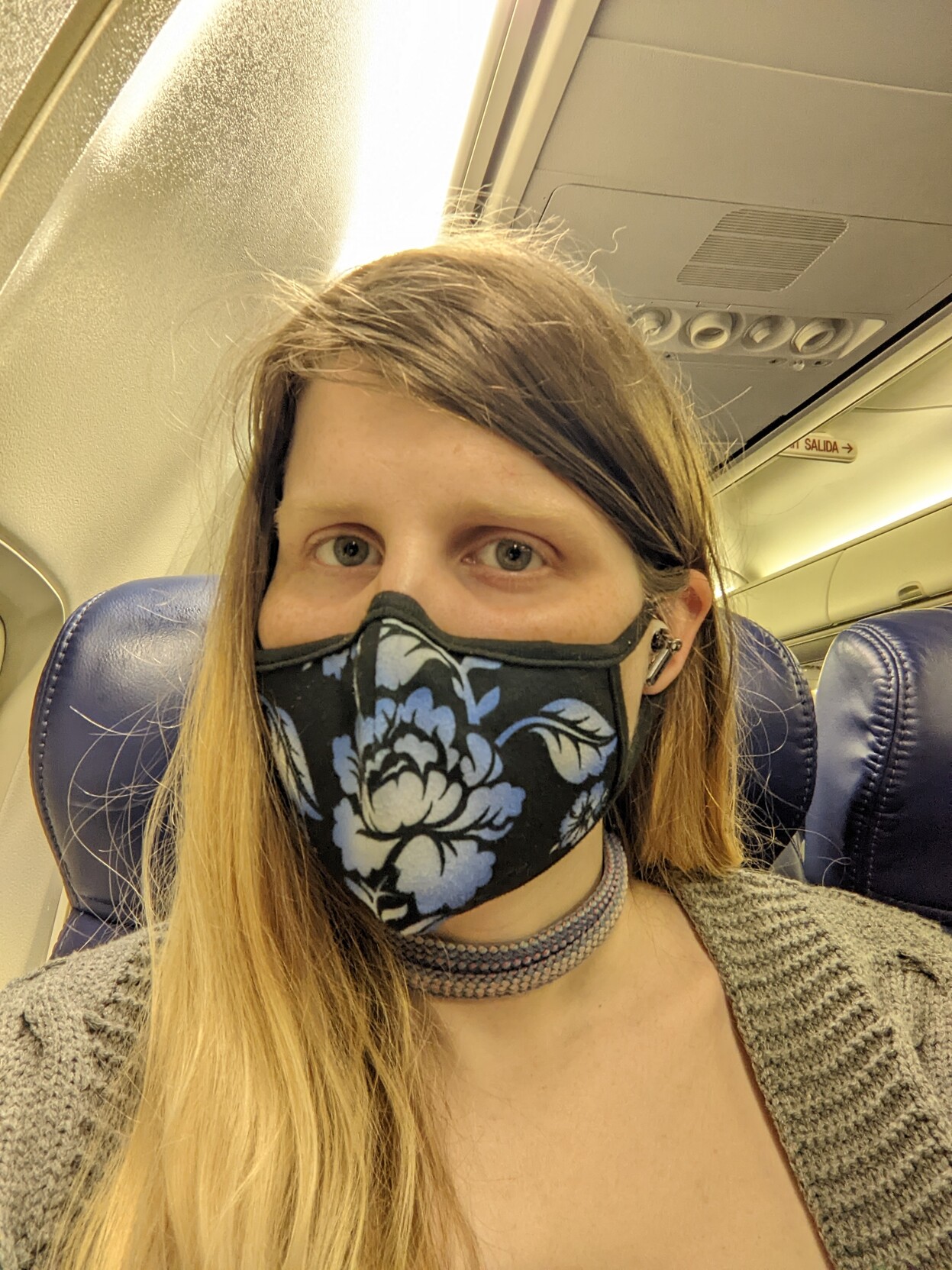 A tired woman on an airplane wearing a black floral print mask and a grey cardigan