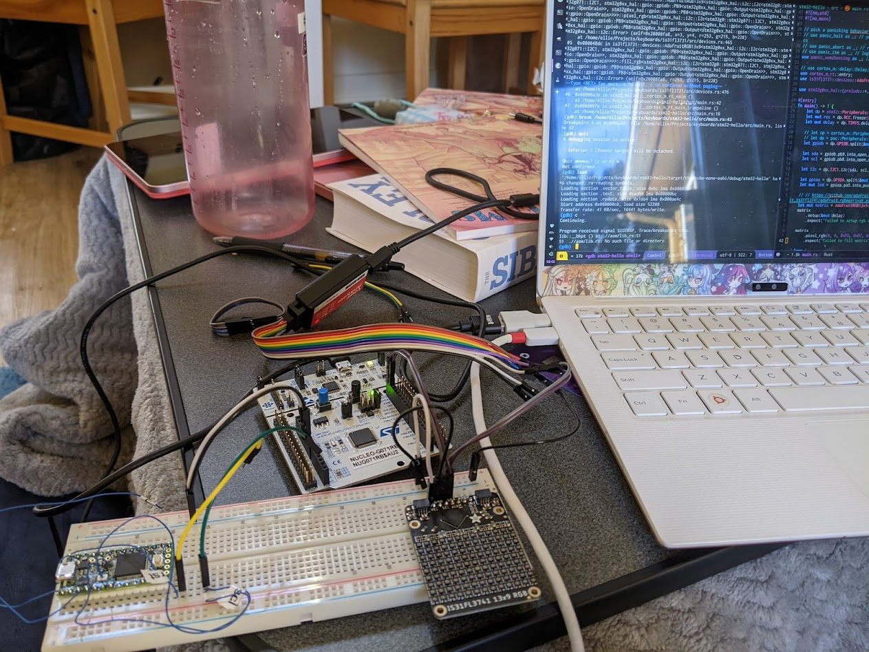 A pile of electronics all wired to each other. To the right, a laptop is running a copy of emacs and debugging the rust code running on the electronics. The table is visibly messy.