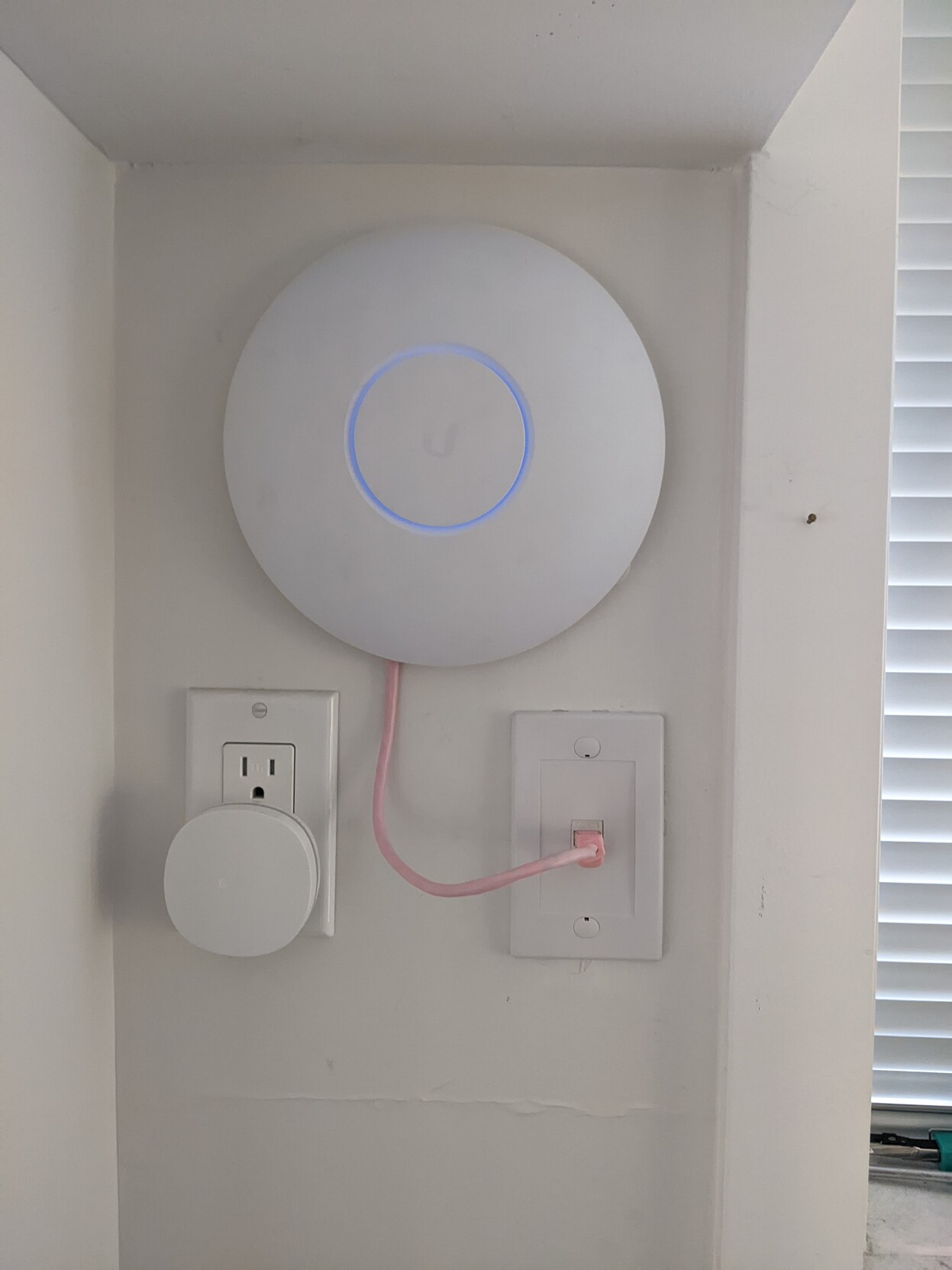 A circular WIFI access point, mounted in a little alcove above an Ethernet port. A pale pink Ethernet cable connects the router to the port