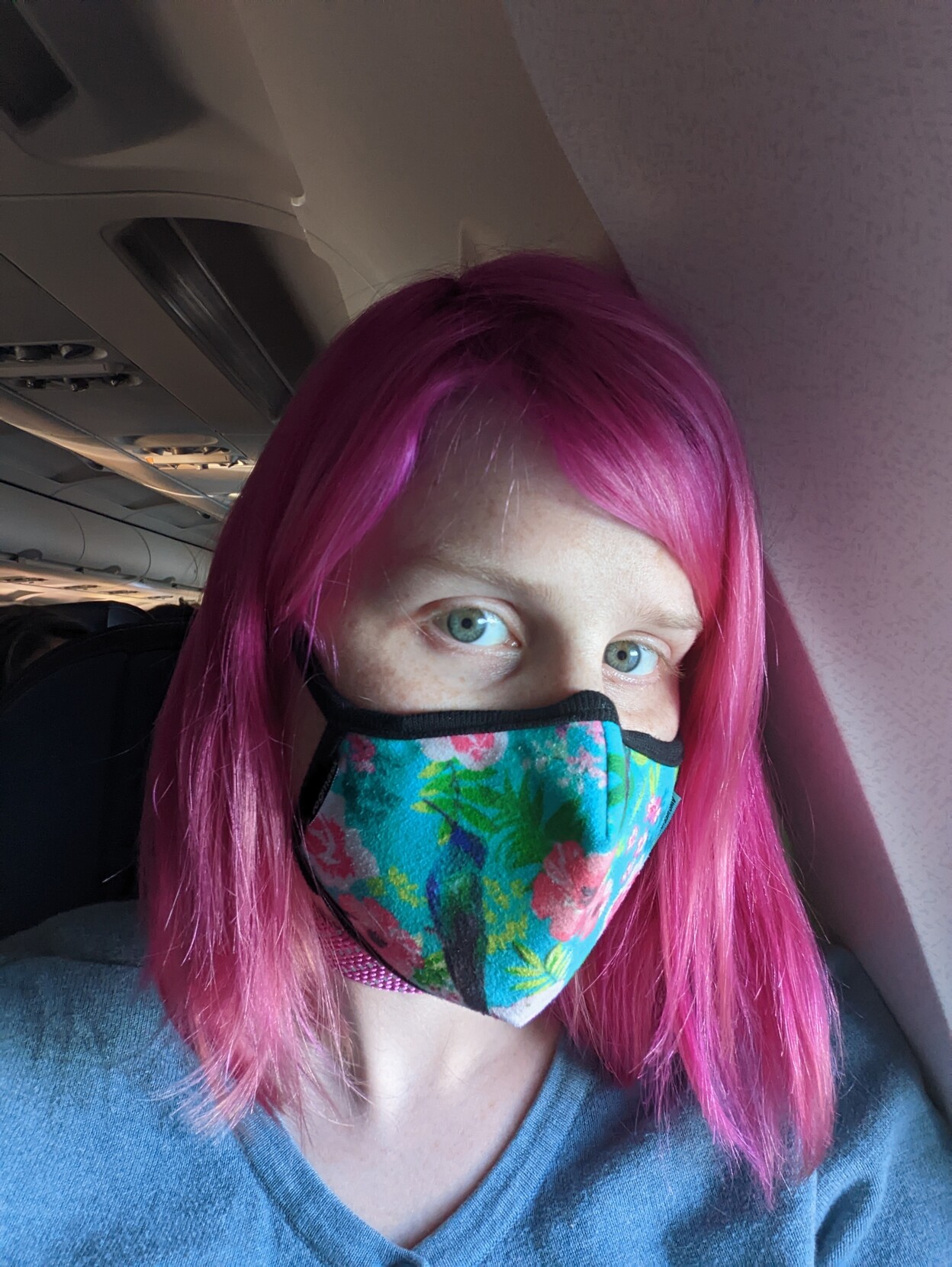 A woman with a pink hair and a colourful blue face mask leaning against the side of an airplane