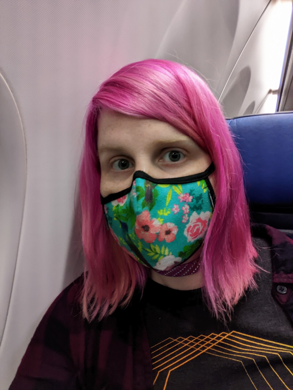 A woman with shiny pink hair, wearing a light blue mask with flowers printed on it, leading against the side of plane