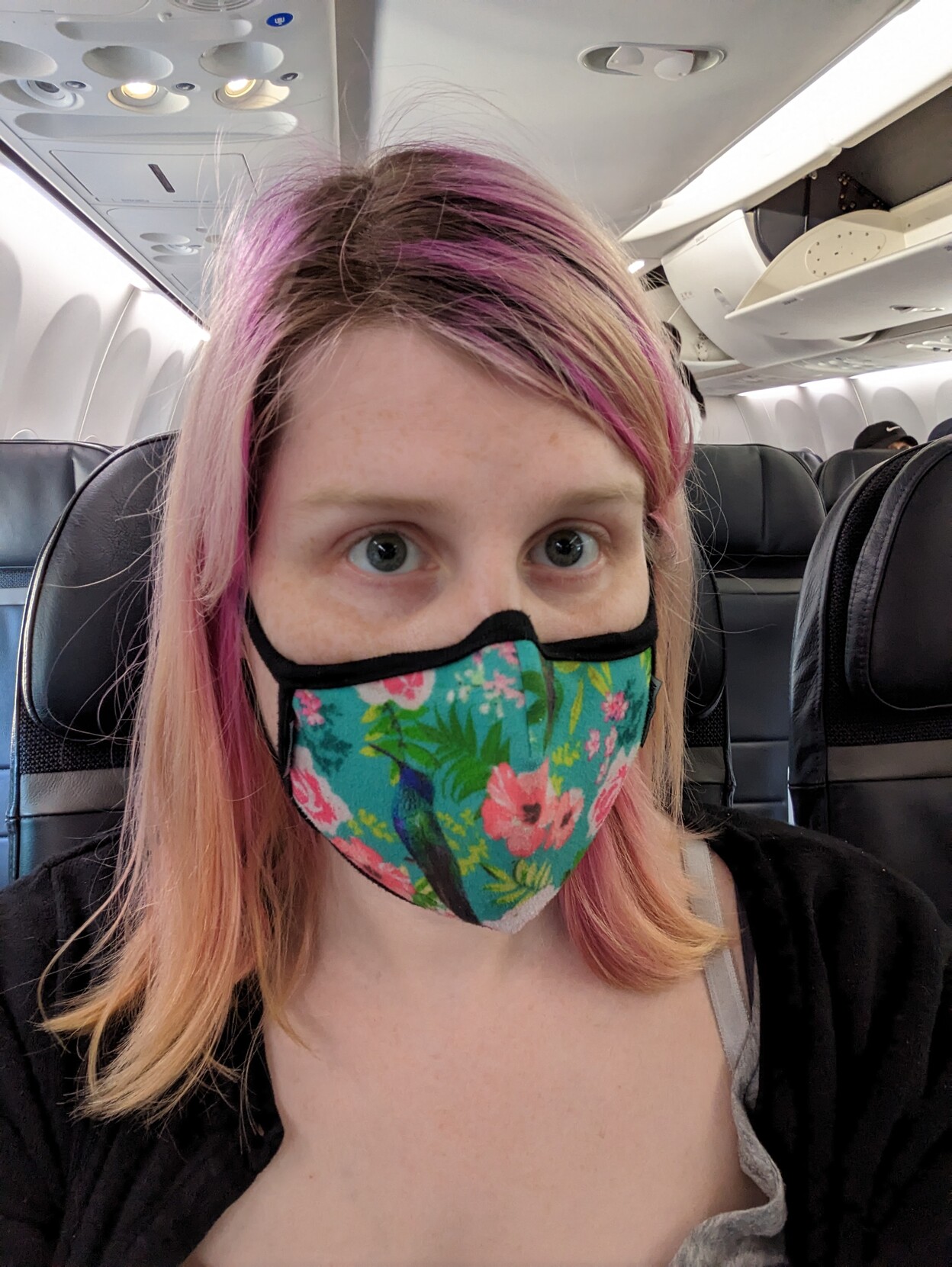 A woman with dyed pink hair and brown routes wearing a teal face mask with prints of birds and flowers 