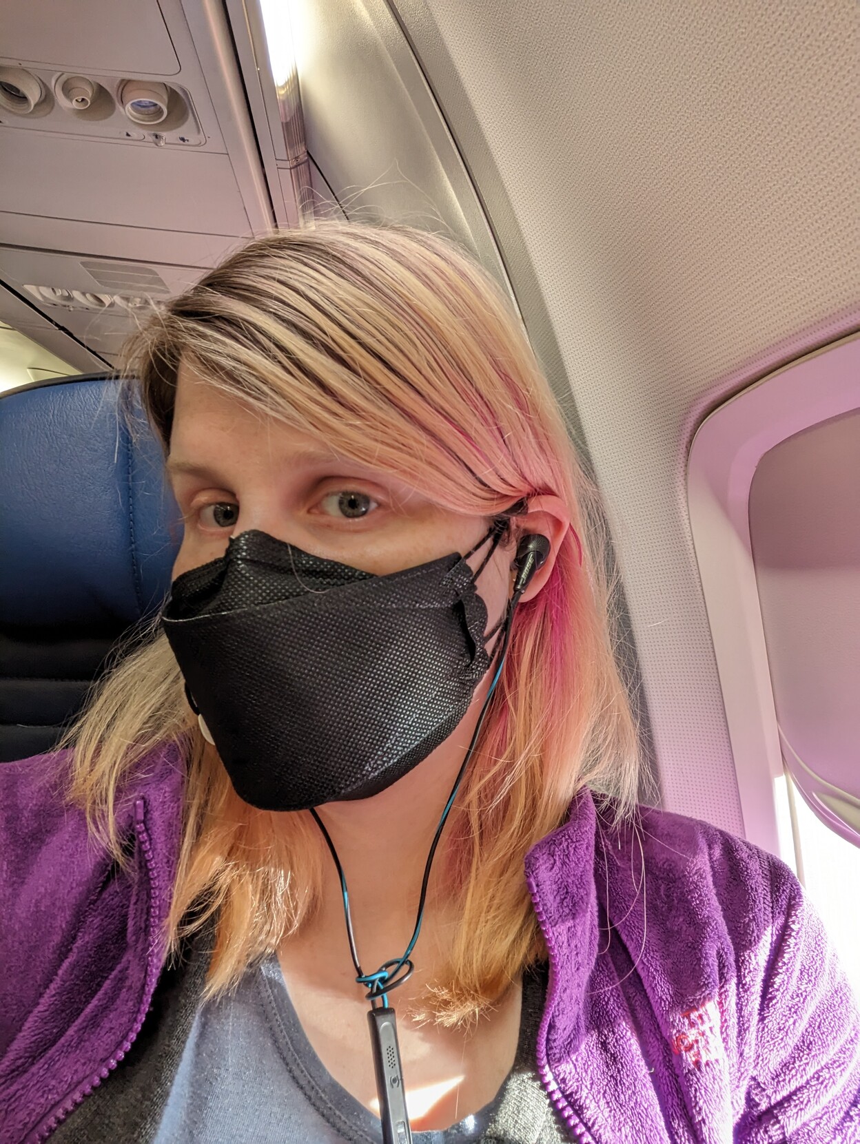 A smiling mostly blond me on a plane wearing a black face mask and purple jacket