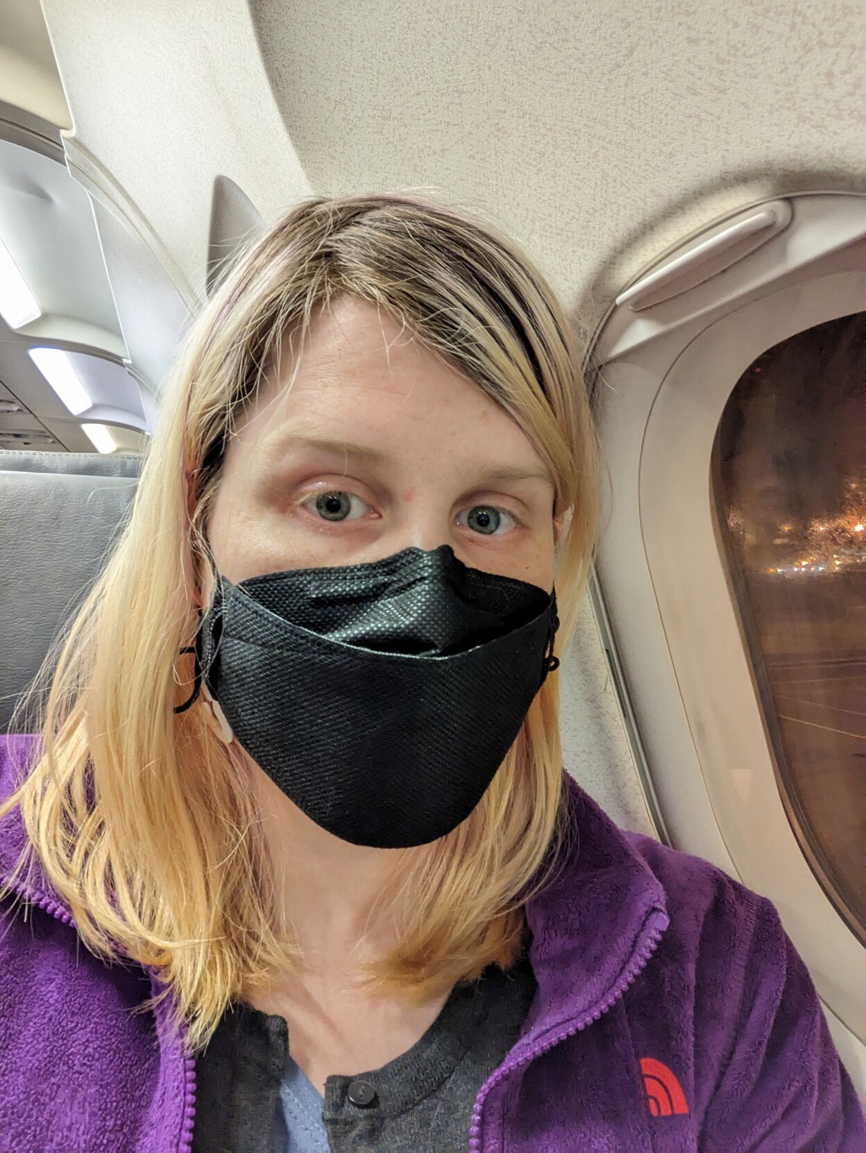 A tired woman (me) on a plane wearing a purple jacket and a black face mask